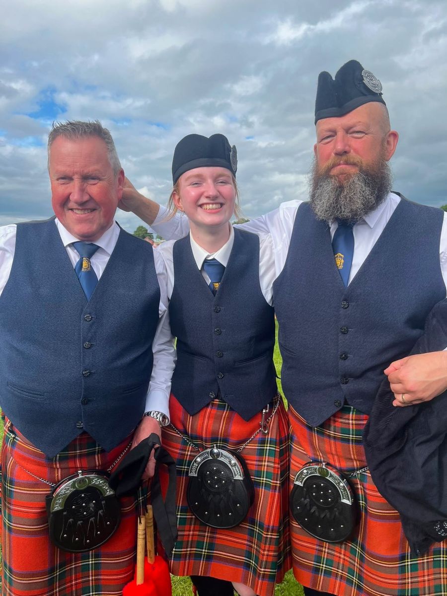 Three band members smiling together in navy waistcoats, ties and red tartan kilts