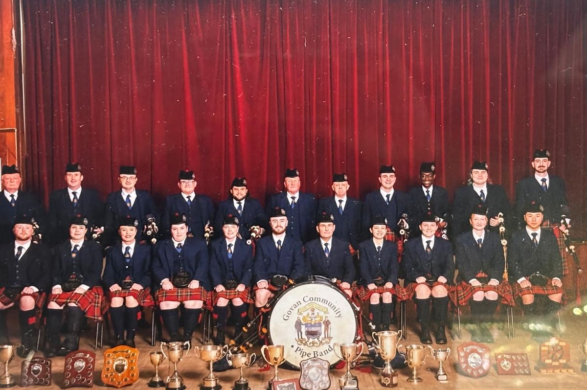 The band seated in formal uniform in front of a red curtain with their trophies at their feet